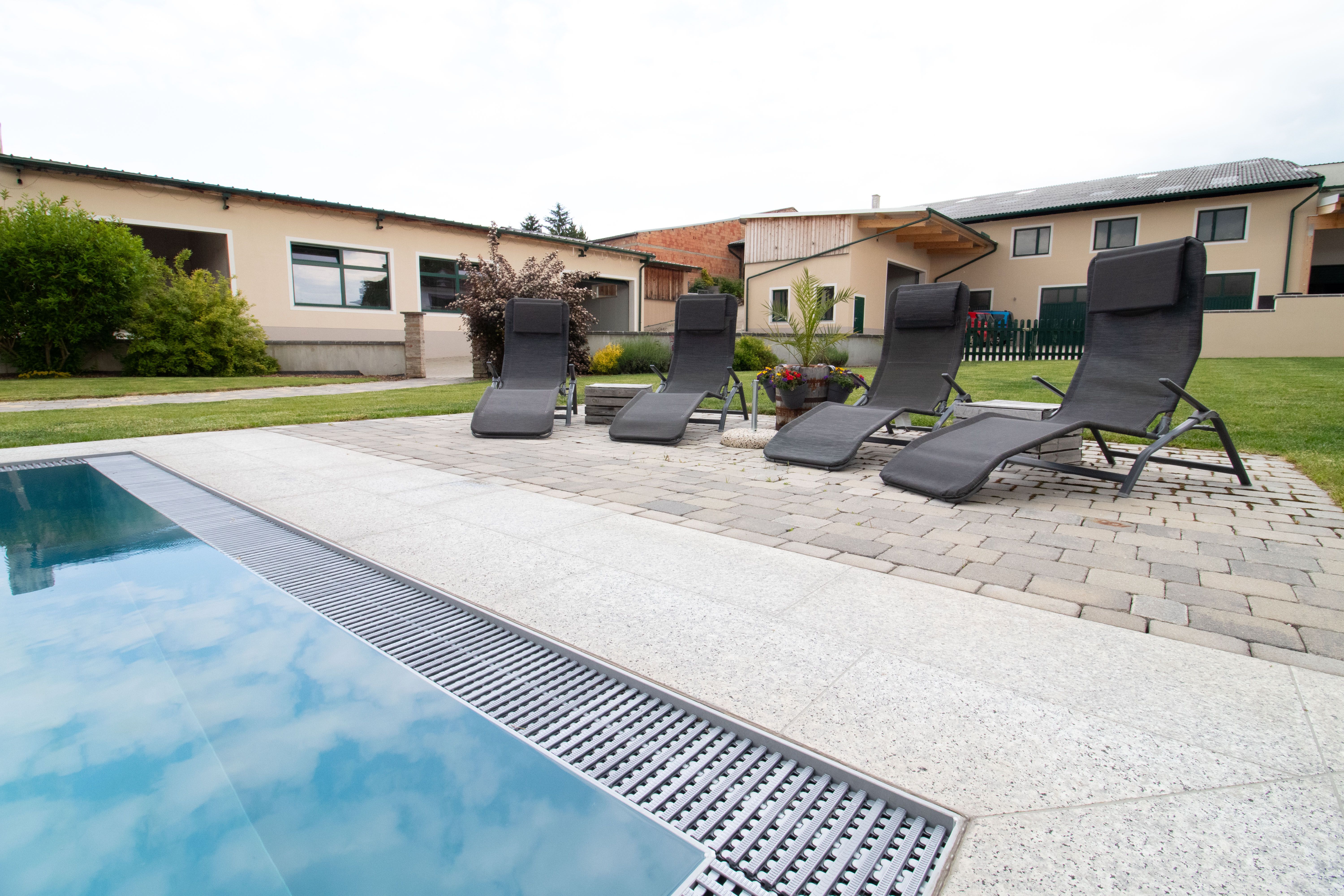 Four lounge chairs are set up next to a pool on a paved terrace. Buildings and a garden can be seen in the background.