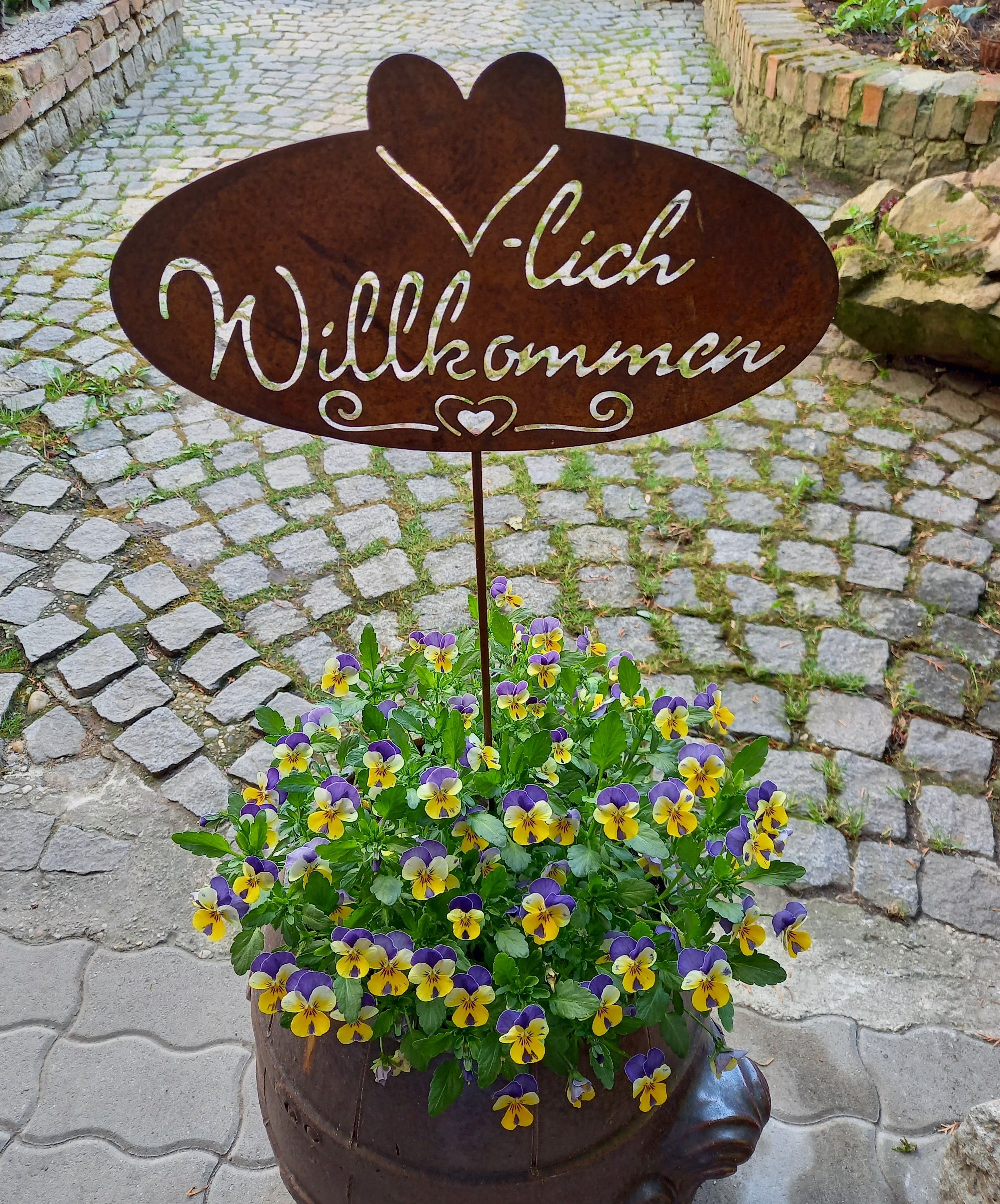 Metal sign with 'Welcome' above a pot of yellow-purple flowers on a cobbled path.