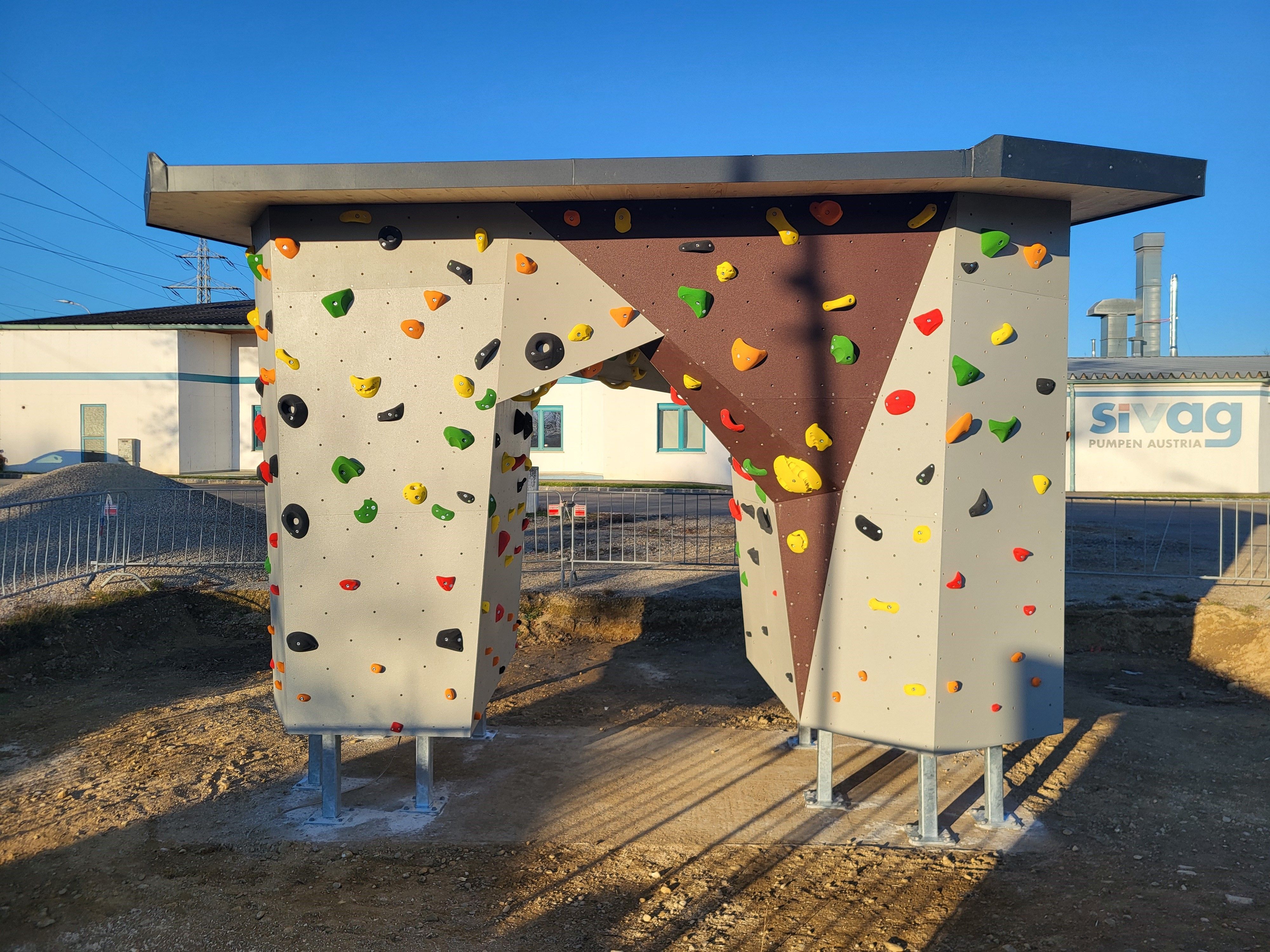 A free-standing bouldering wall with colorful climbing holds on a sunny site.