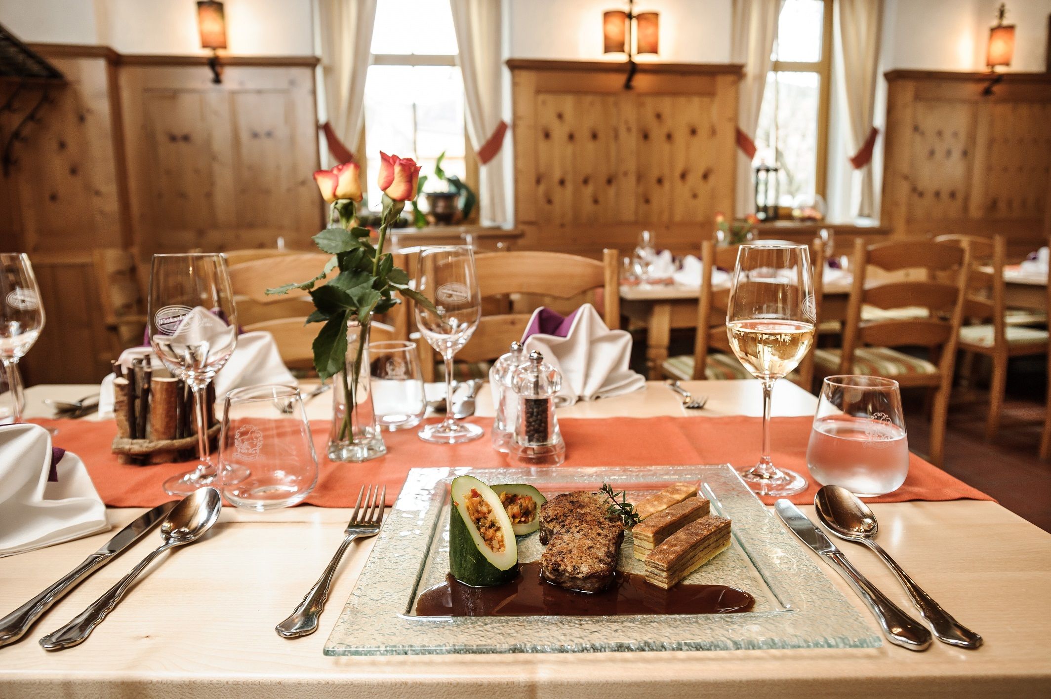 An elegantly laid table in a rustic restaurant with a plate of gourmet food, wine glasses and a vase of roses.