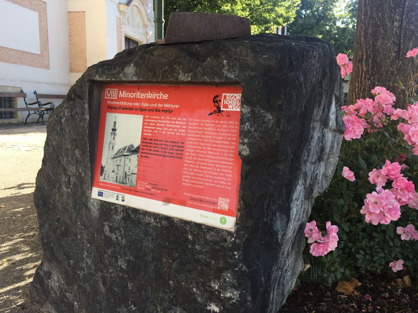 Stone plaque with information about the Minorite Church, surrounded by flowers.