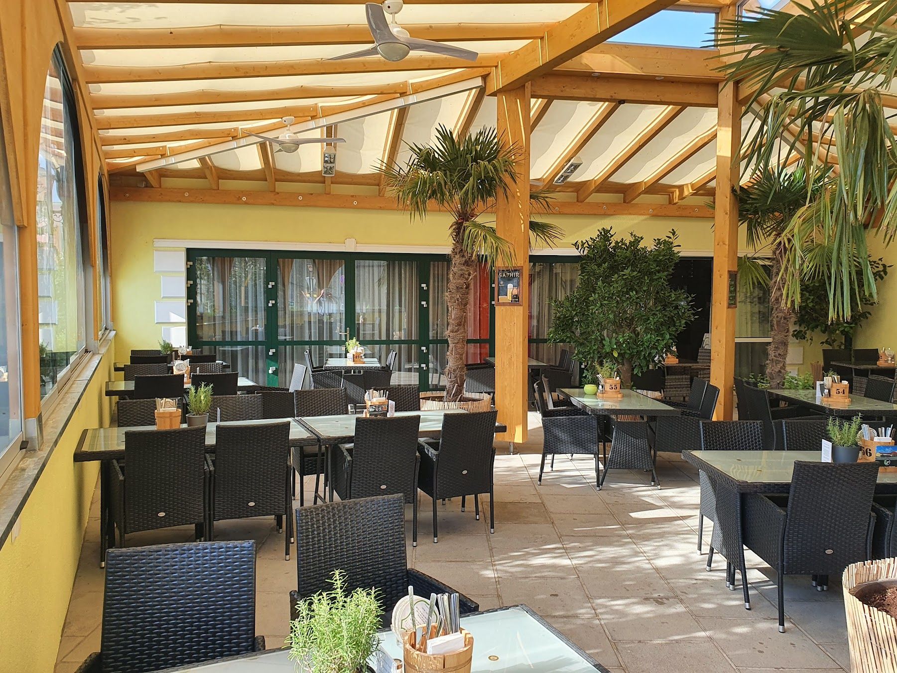 Covered terrace of a restaurant with black rattan furniture and plants.