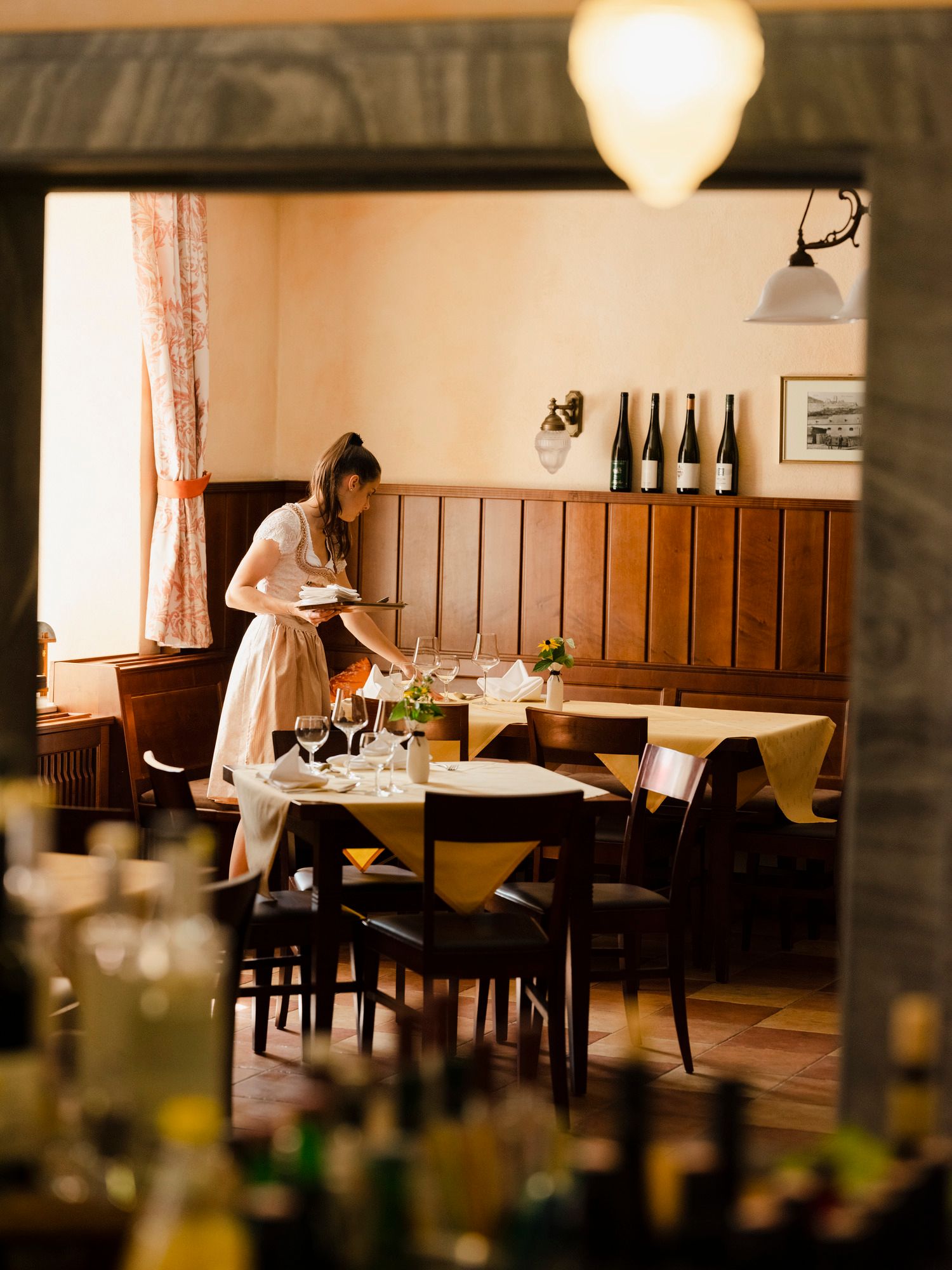 A woman in traditional dress sets a table in a cozy restaurant with wooden walls and bottles of wine in the background.