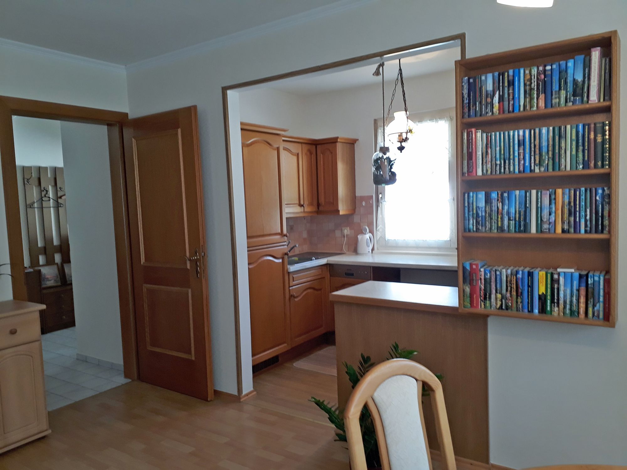 Interior view of a kitchen with wooden cupboards, a bookshelf and a dining table.