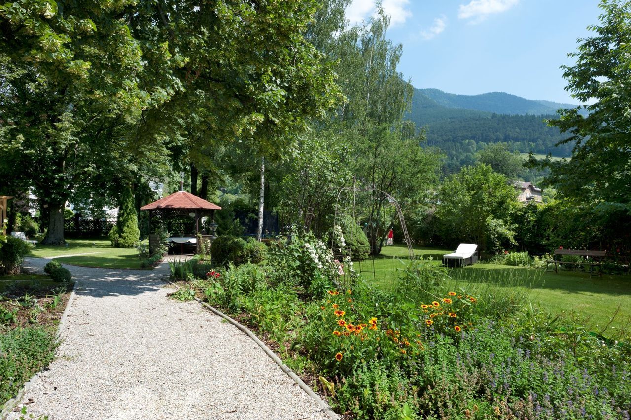 A well-kept garden with a gravel path, flower beds, a pavilion and a lounger. Trees and mountains can be seen in the background.