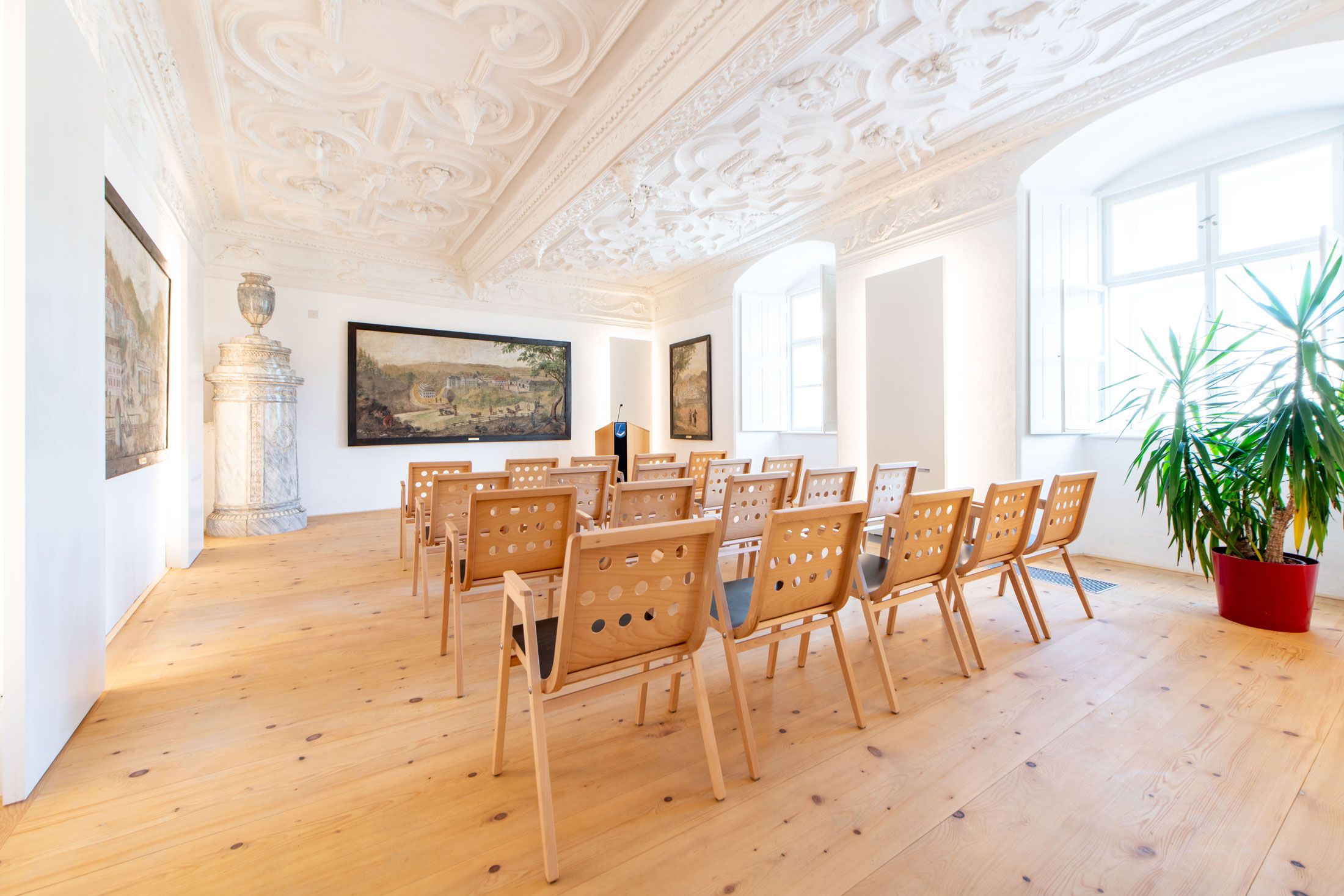 An elegant room with ornate ceilings, wooden floors and rows of chairs facing a screen.