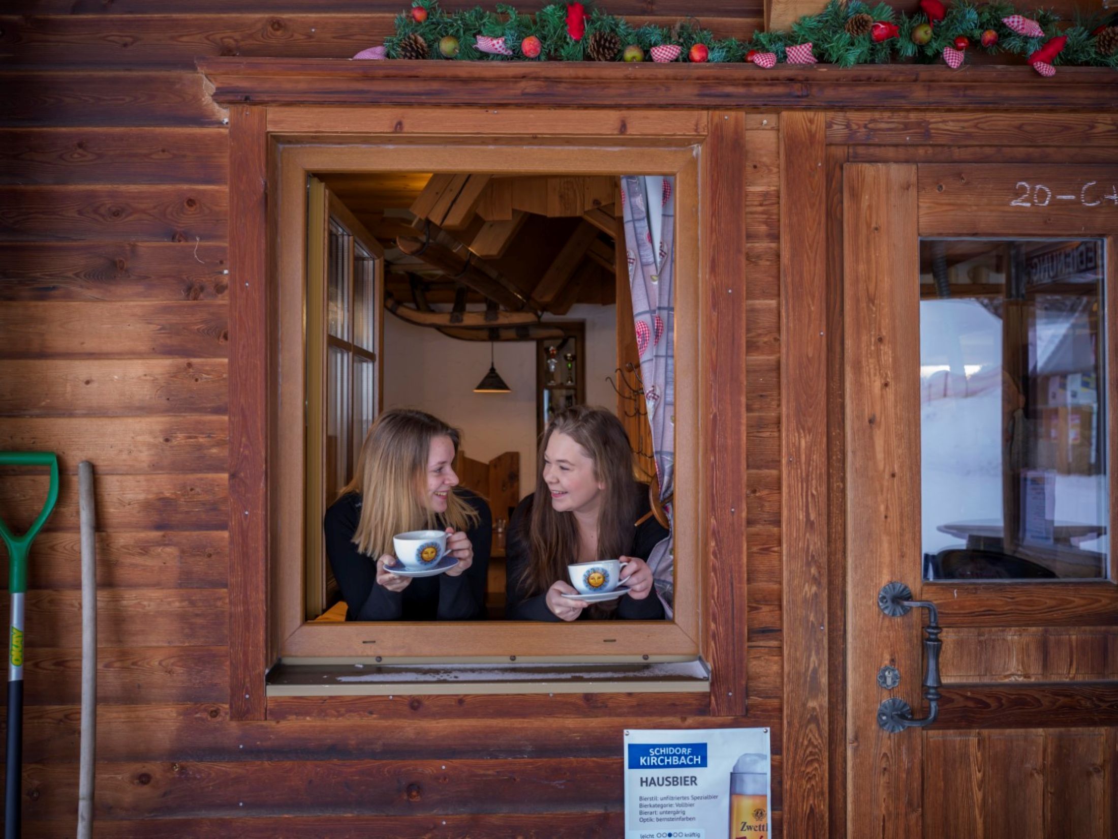 Two women look out of a window of a wooden hut and hold cups in their hands.