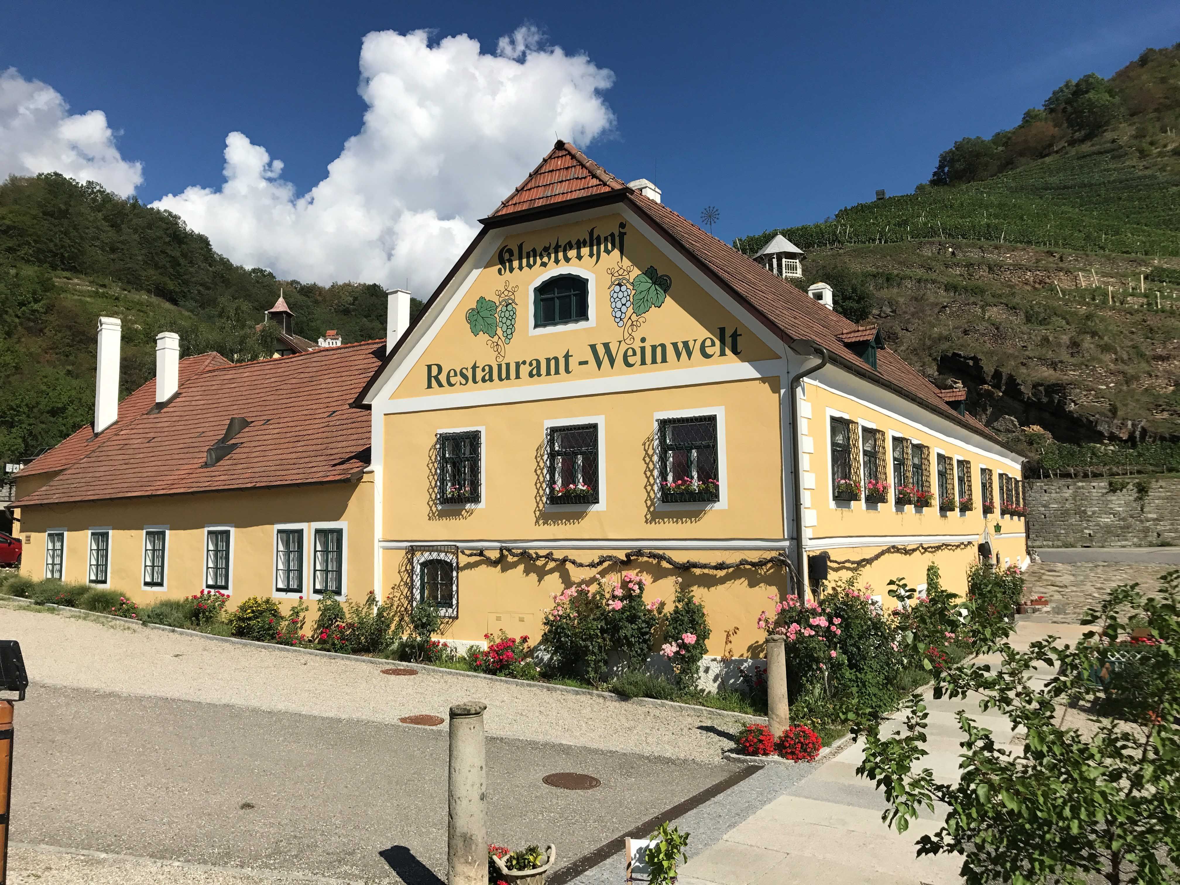 Exterior view of a yellow building with the inscription 'Klosterhof Restaurant-Weinwelt', surrounded by flowers and vineyards.