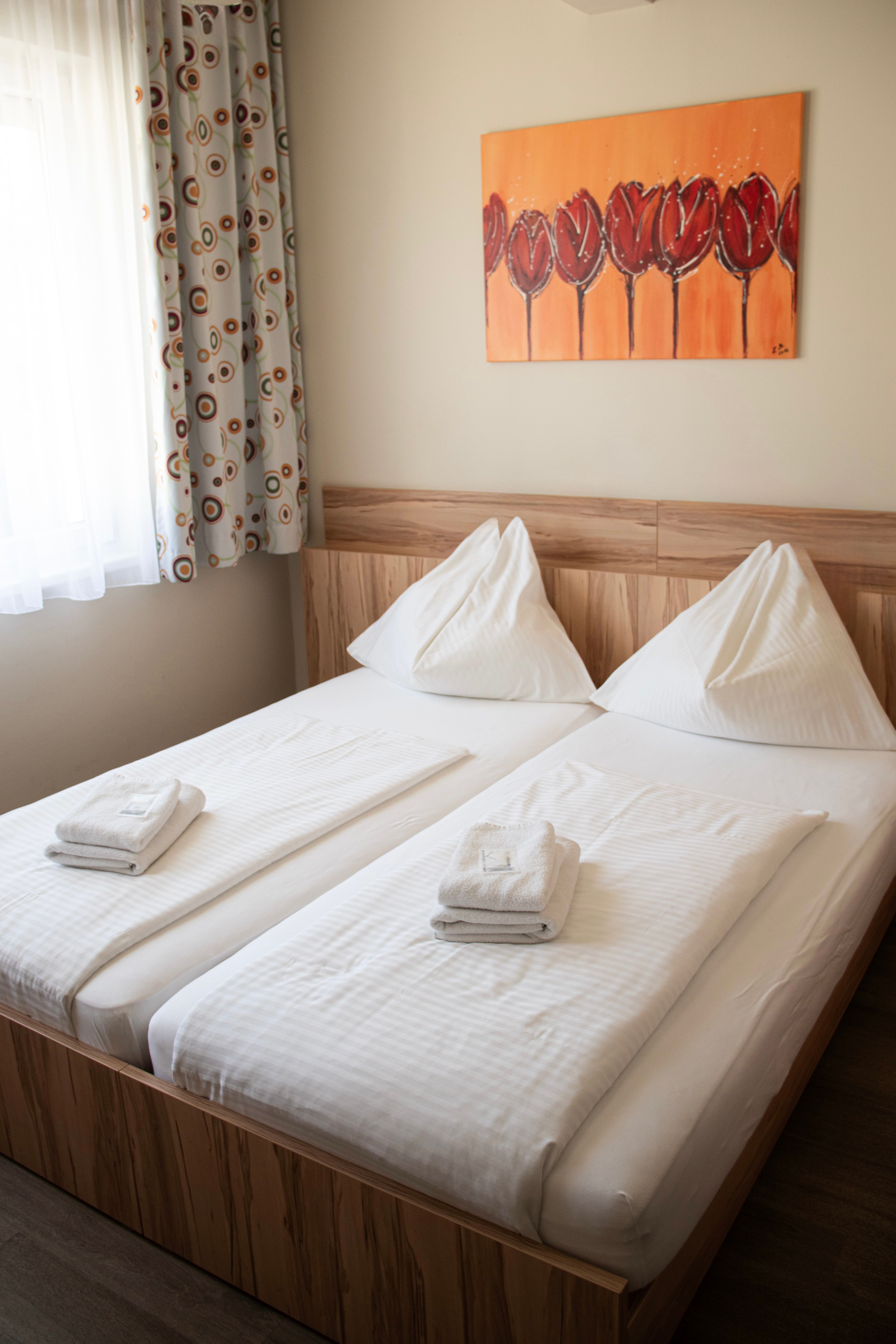 A double bed in the Kolping House with white bedding, towels, a window with patterned curtains, and a wall hanging featuring red tulips.