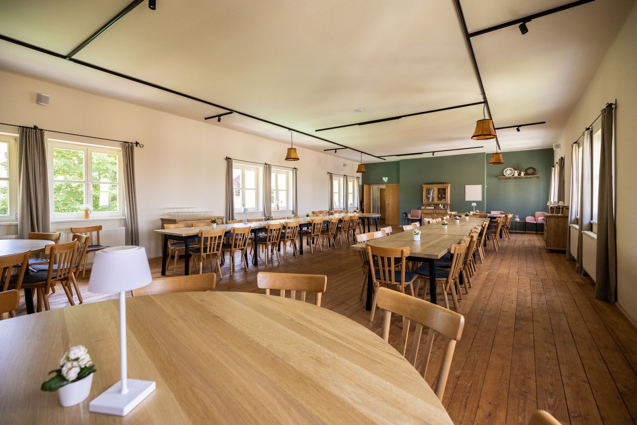 A large, bright room with wooden floors and wooden tables and chairs. Windows on the left wall let in daylight. The room has a modern design, with one wall painted green and the rest white.
