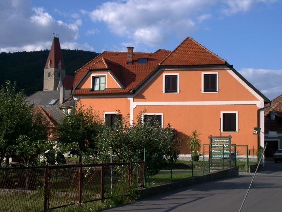 An orange-colored house with red roof tiles and a church tower in the background.