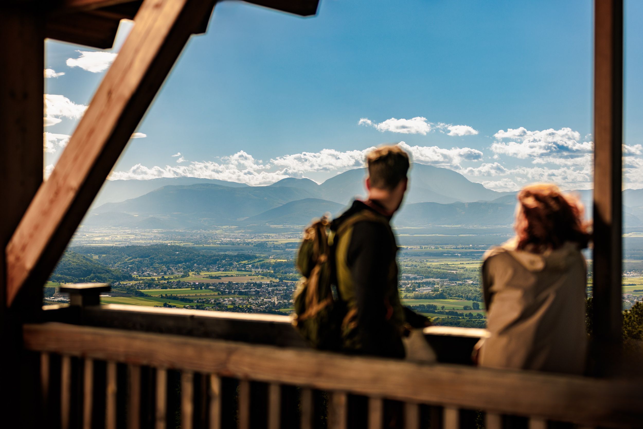 Two people are standing on the platform of the wooden observation tower in Lanzenkirchen/Wiesen, with a wide plain in the background and the Schneeberg clearly visible on the horizon.