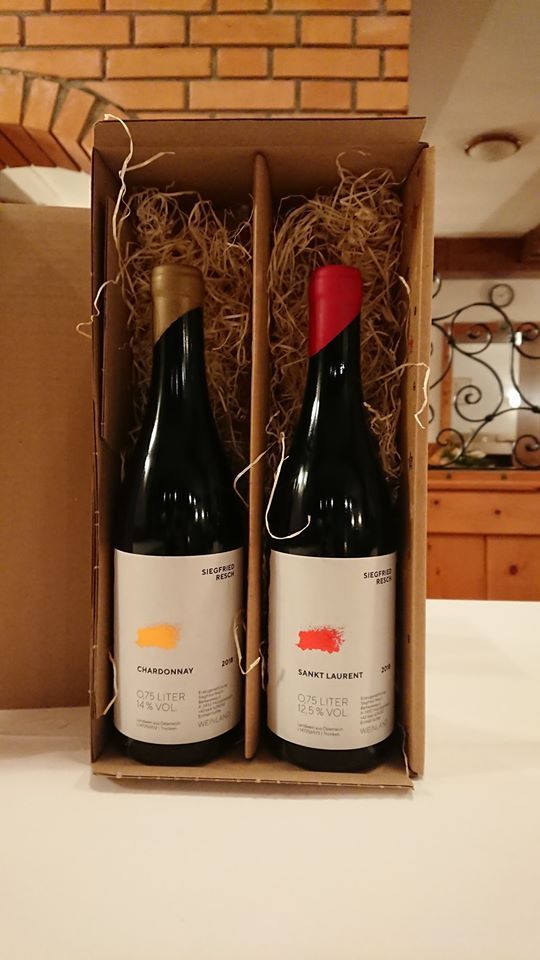 Two bottles of wine in a cardboard box with straw, a brick wall in the background.