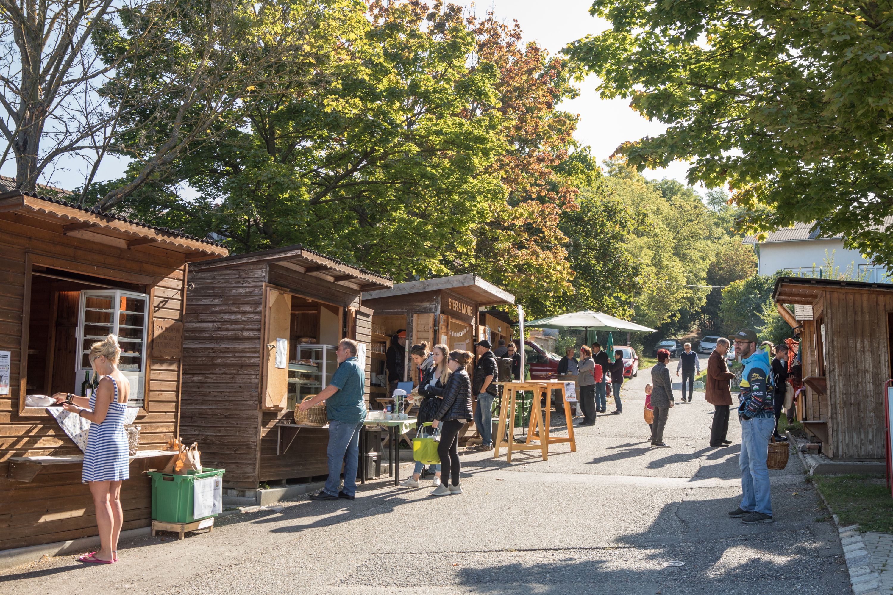 People standing by wooden huts at an outdoor market in sunny weather.