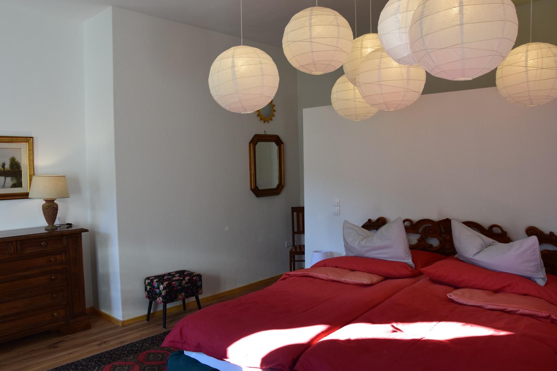 Bedroom with double bed, red comforters, wooden chest of drawers, lamp, mirror and paper lamps on the ceiling.