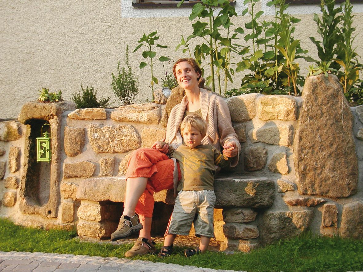 A woman and a child sit on a stone bench in front of a rock garden overgrown with plants.