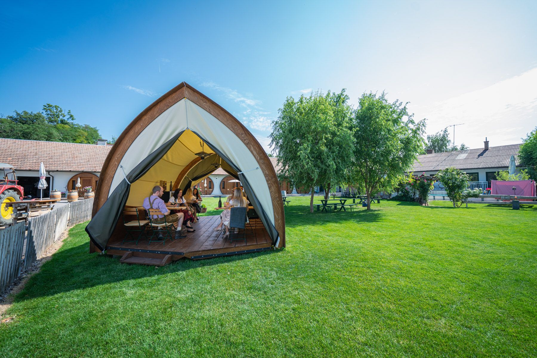 People sit in an open tent on a meadow, surrounded by trees and buildings.