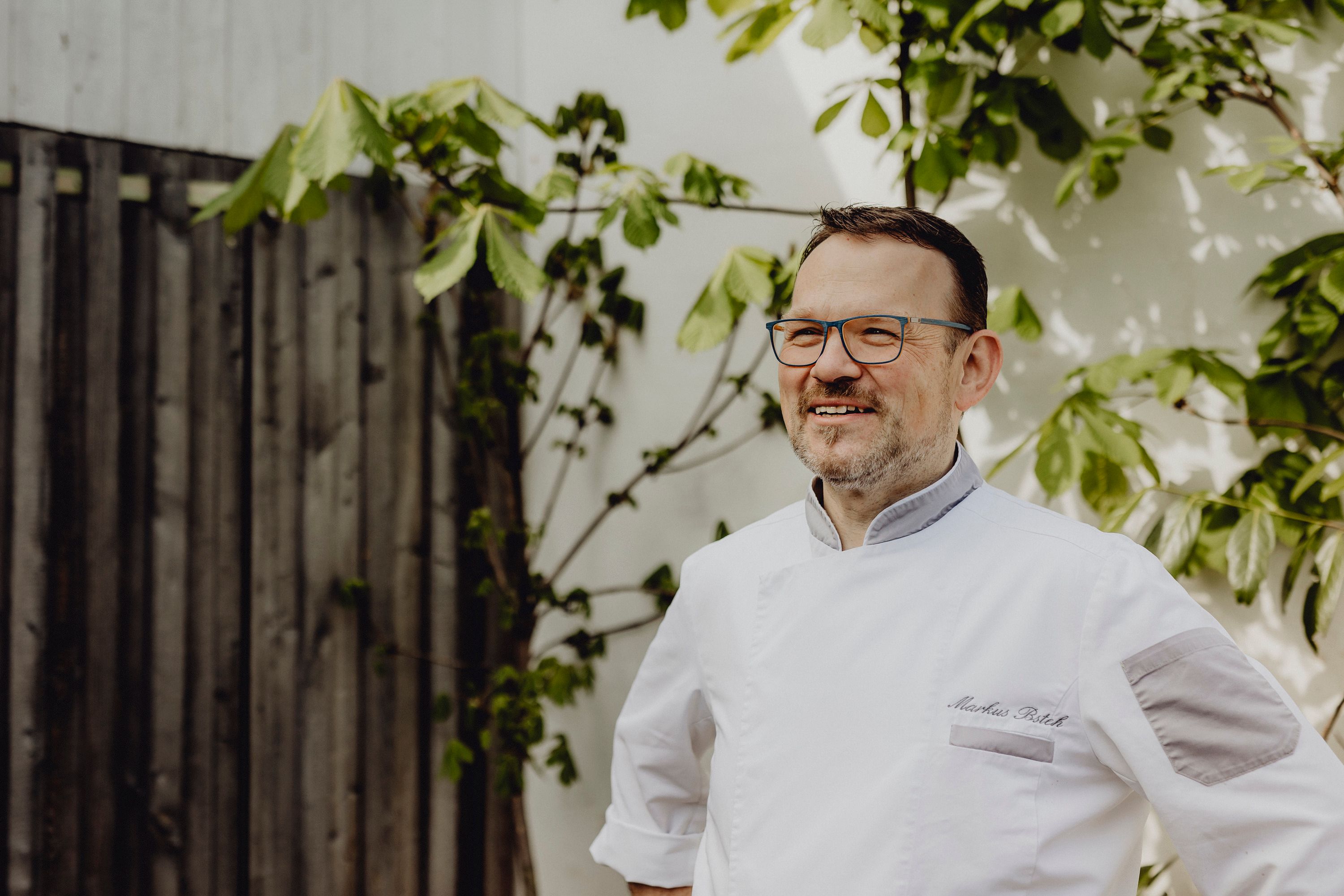 A man in chef's clothes stands smiling in front of a wooden wall with plants.
