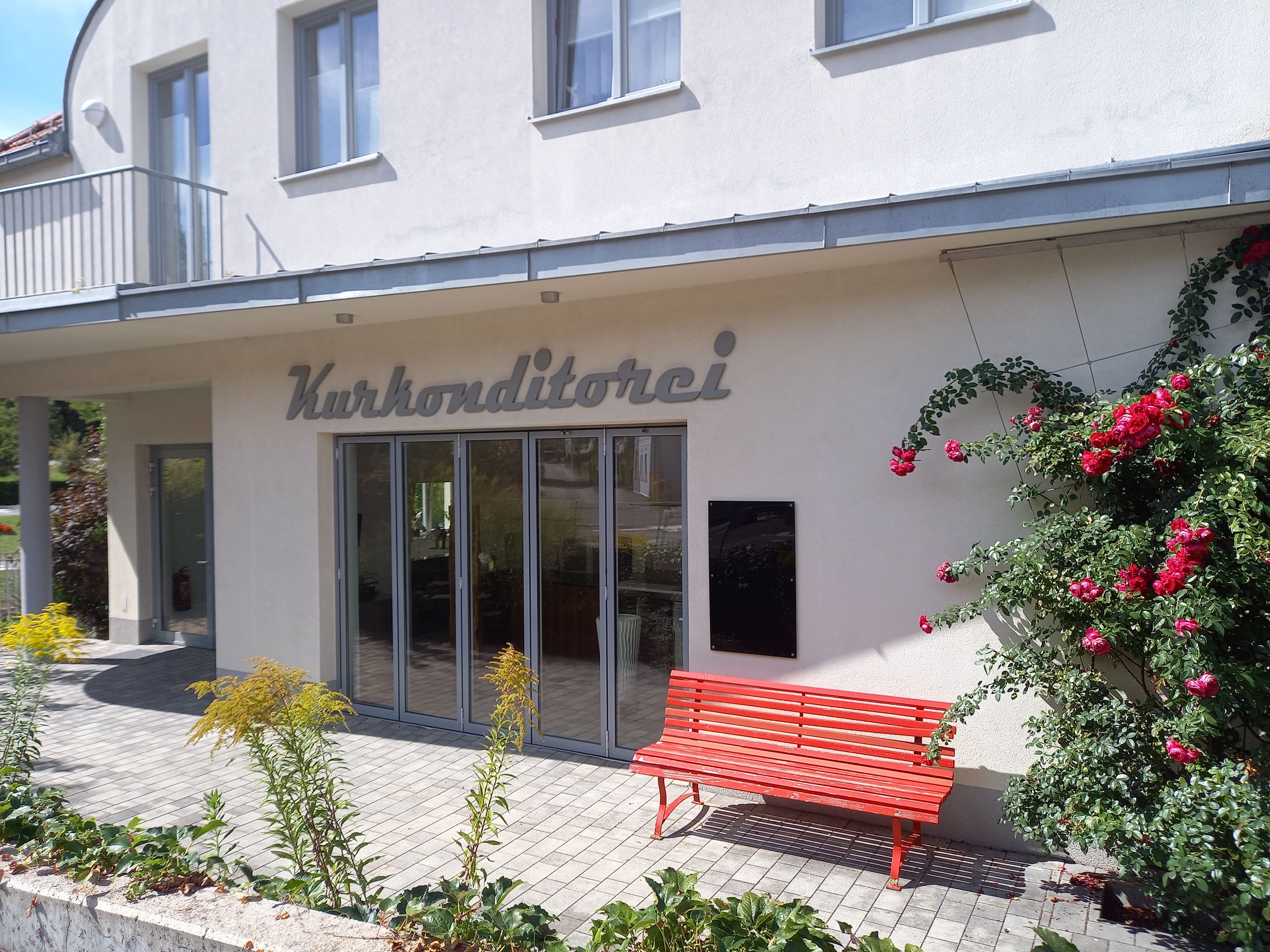 Exterior view of a building with the inscription 'Kurkonditorei', red bench and blooming roses.