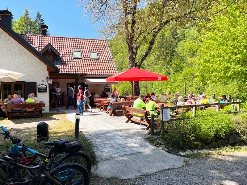 A mountain hut with a terrace where people are sitting at tables. Bicycles can be seen in the foreground, surrounded by green nature.