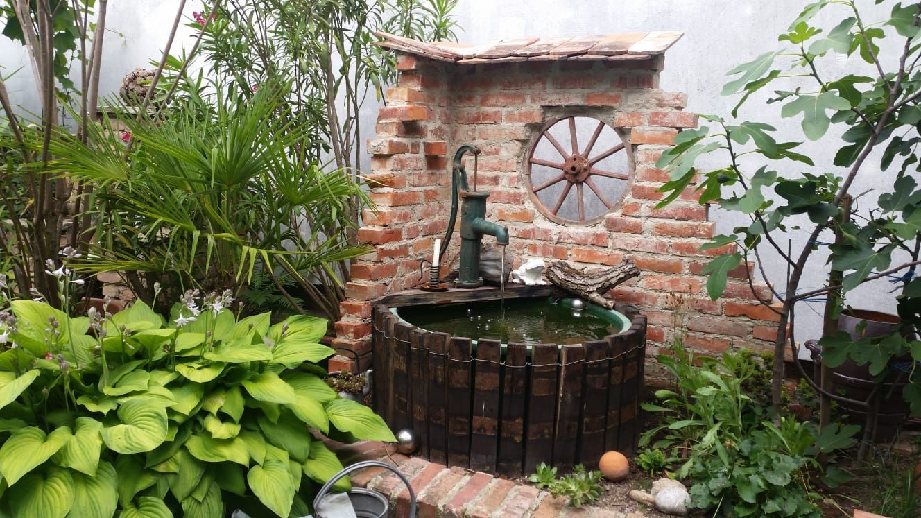 A rustic garden fountain with a brick wall, surrounded by plants and an old wagon wheel as decoration.
