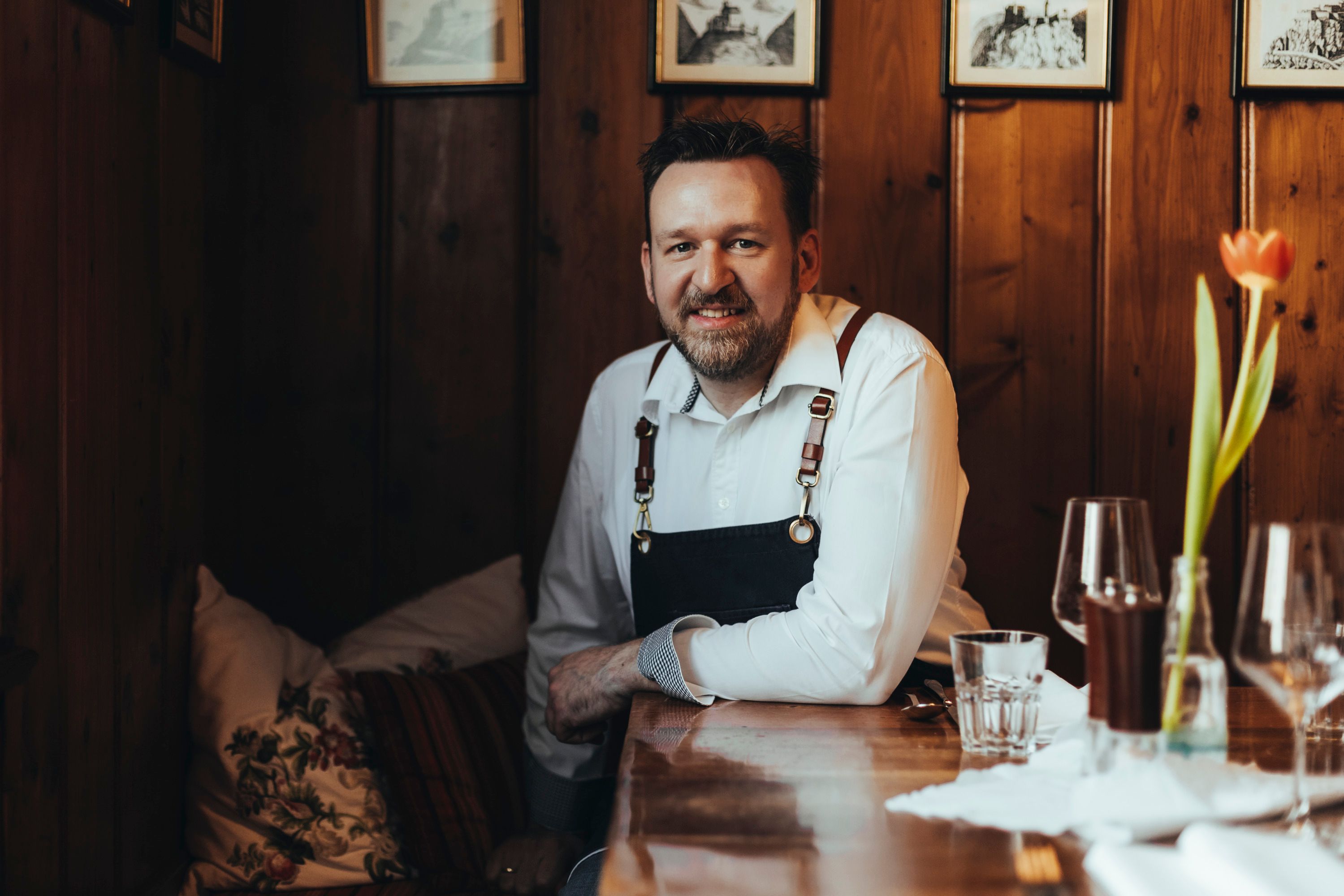 A man in a restaurant is sitting at a wooden table, wearing a white shirt and an apron. In the foreground is a vase with a tulip.