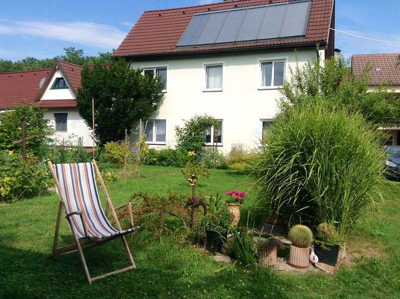 A house with solar panels on the roof, surrounded by a garden with plants and a sun lounger.
