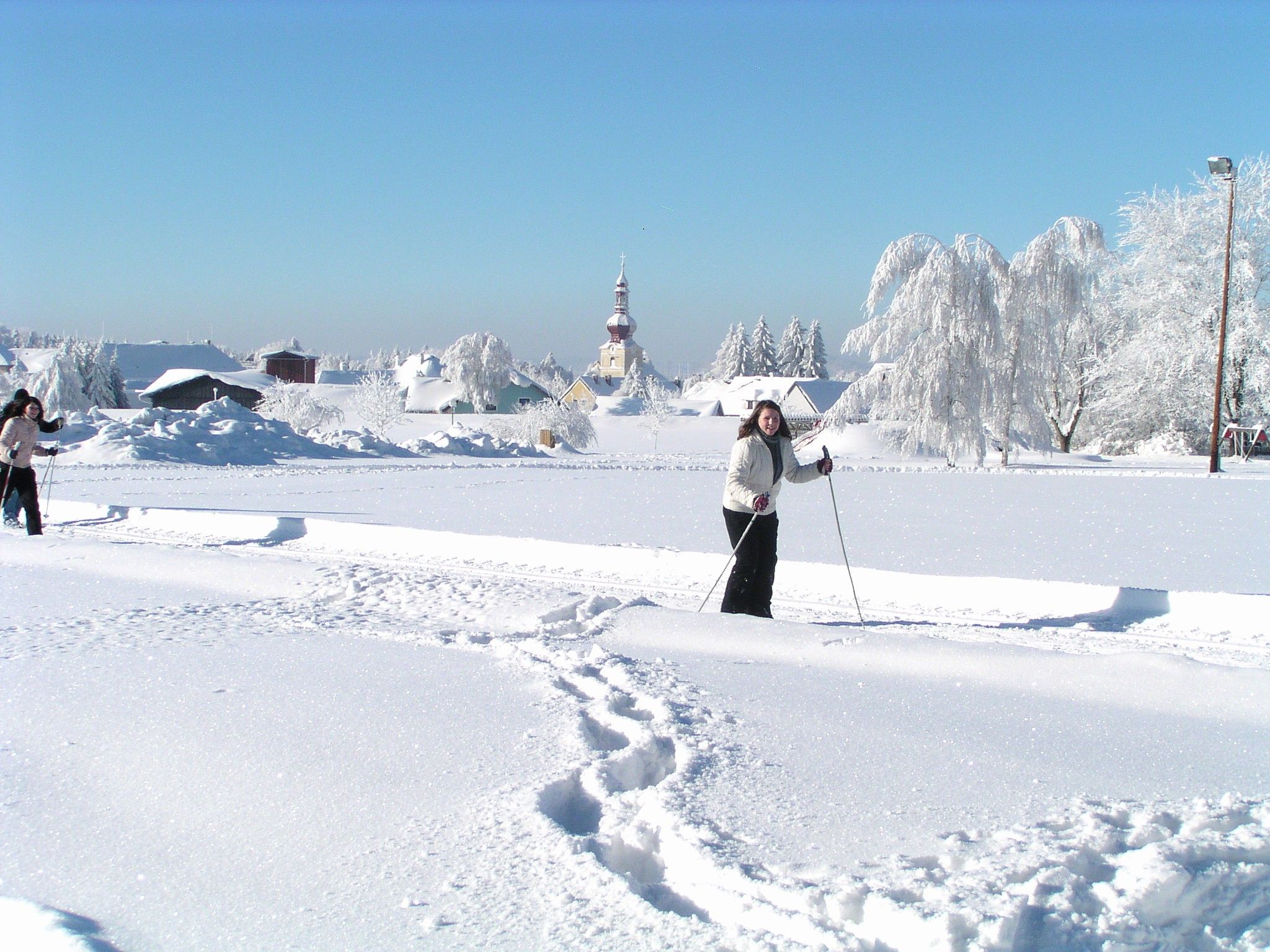 Two people cross-country skiing in a snowy landscape with a church in the background.