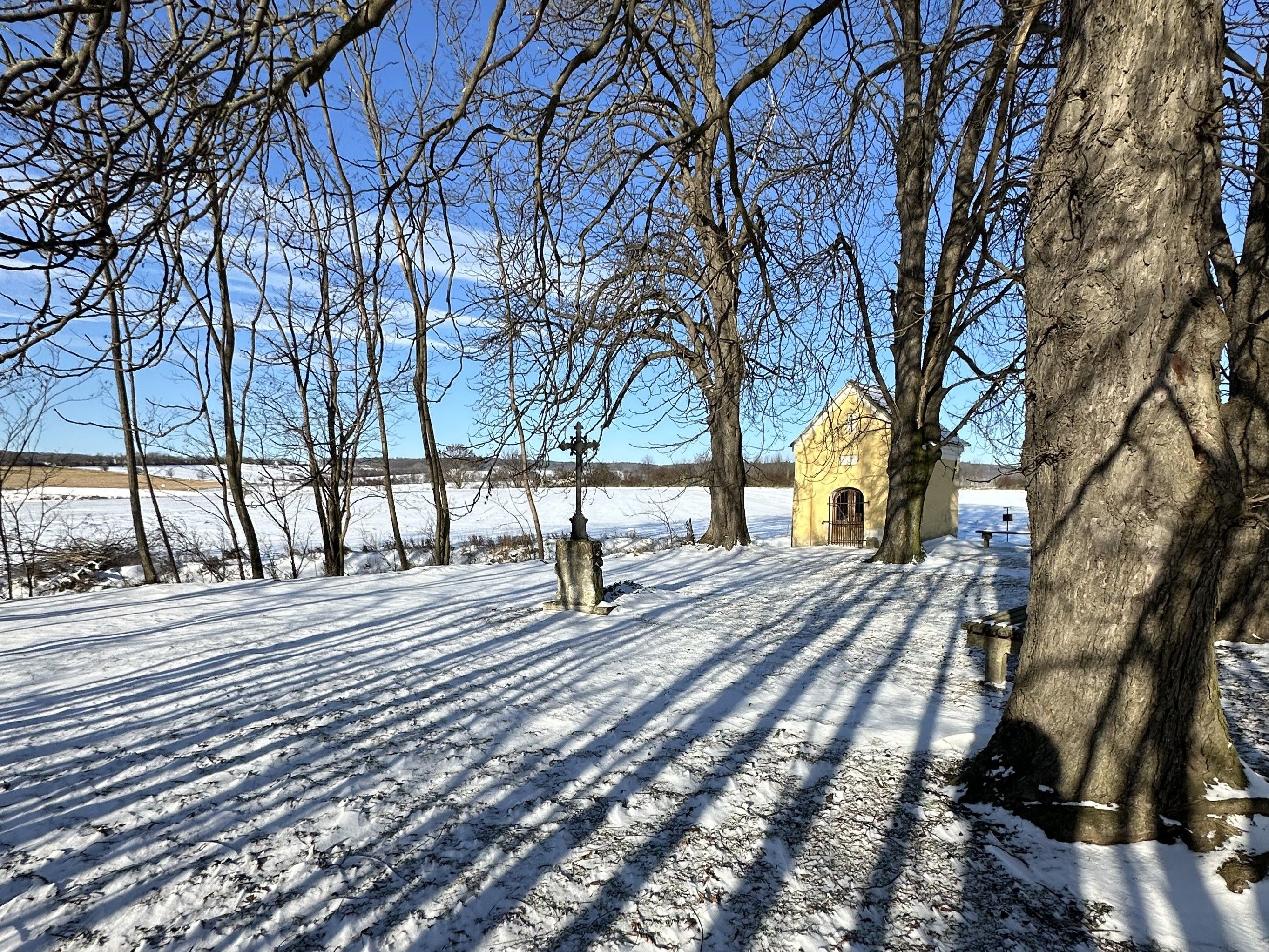 Winter landscape with a small yellow chapel in the background, surrounded by snow-covered trees and fields.