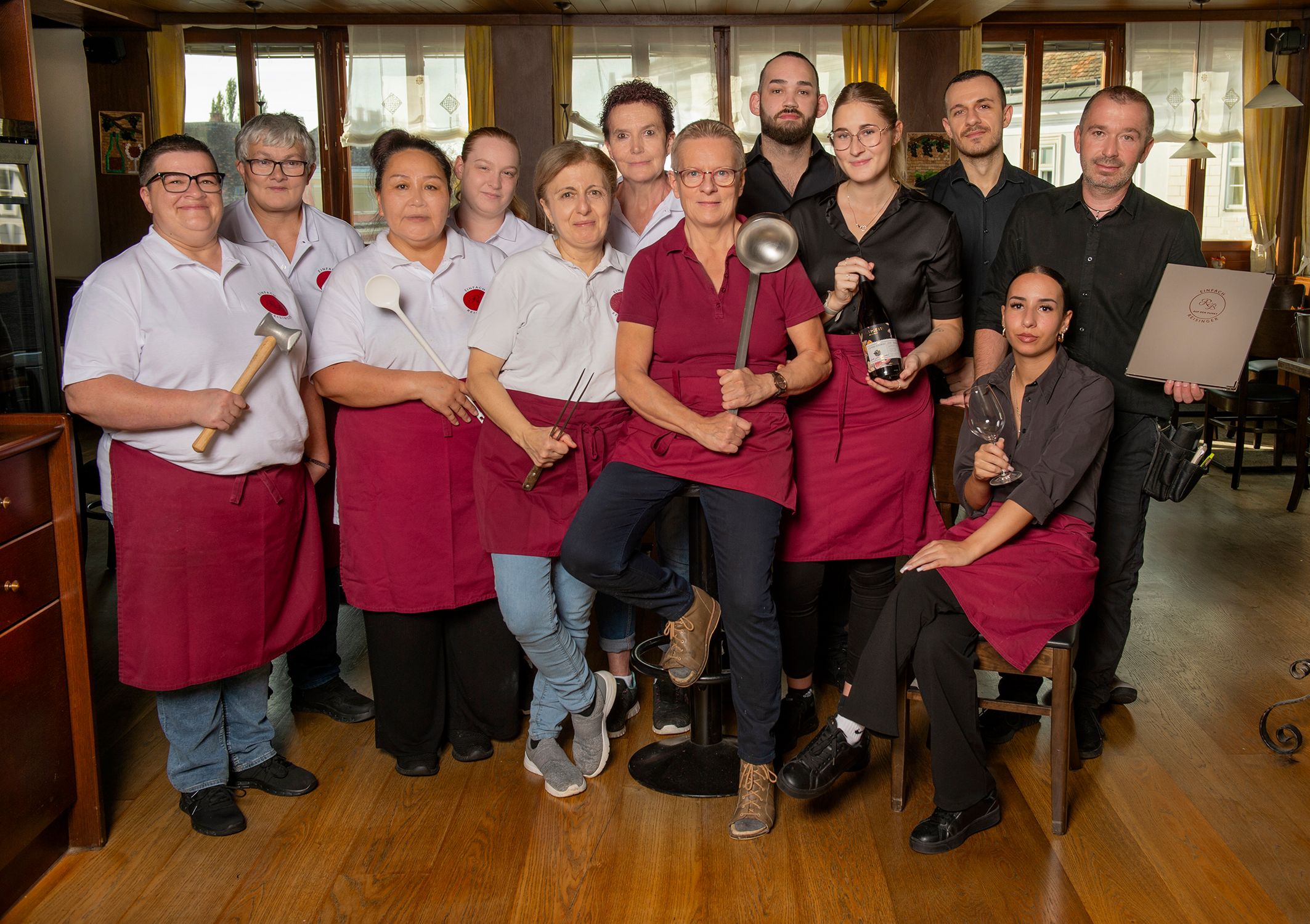A team of restaurant staff pose in work clothes with kitchen utensils in a dining room.