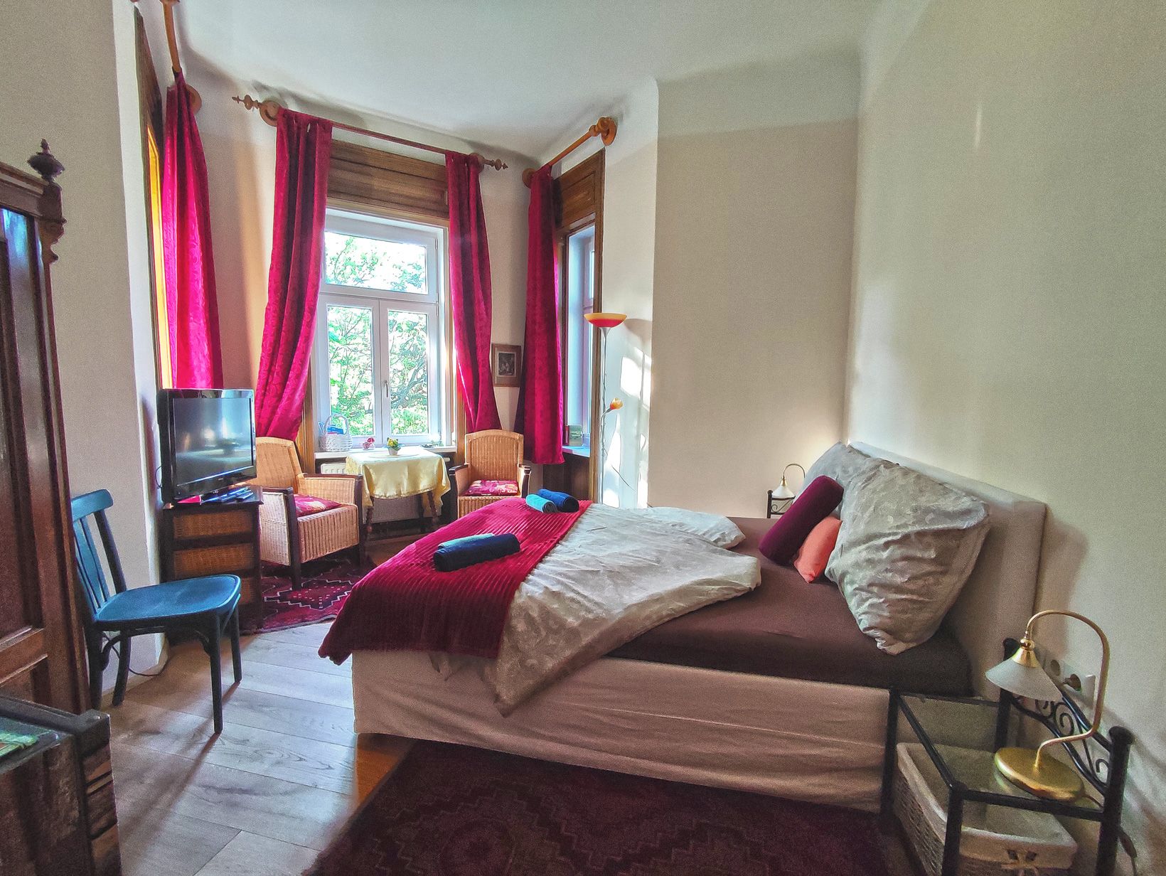 Cozy bedroom with double bed, red curtains and armchairs in front of a window.