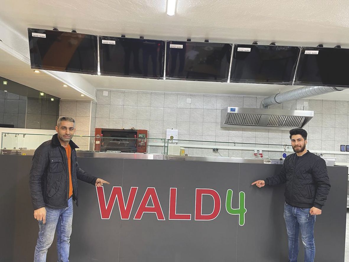Two men are standing in front of a counter labeled 'WALD4' in a pizzeria.
