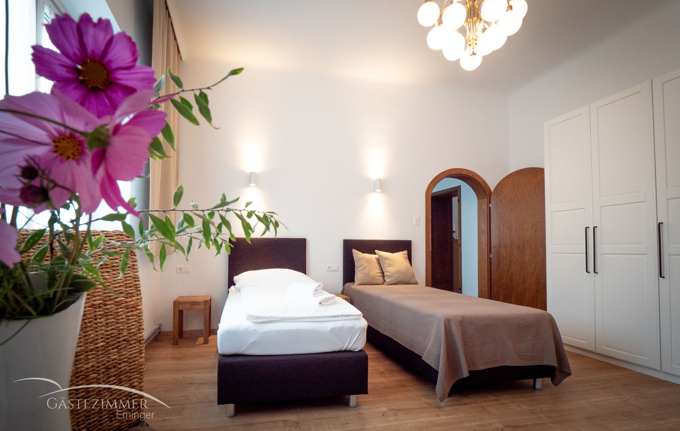 A cozy twin room with flowers in the foreground, two beds, a wardrobe and a lamp on the ceiling.