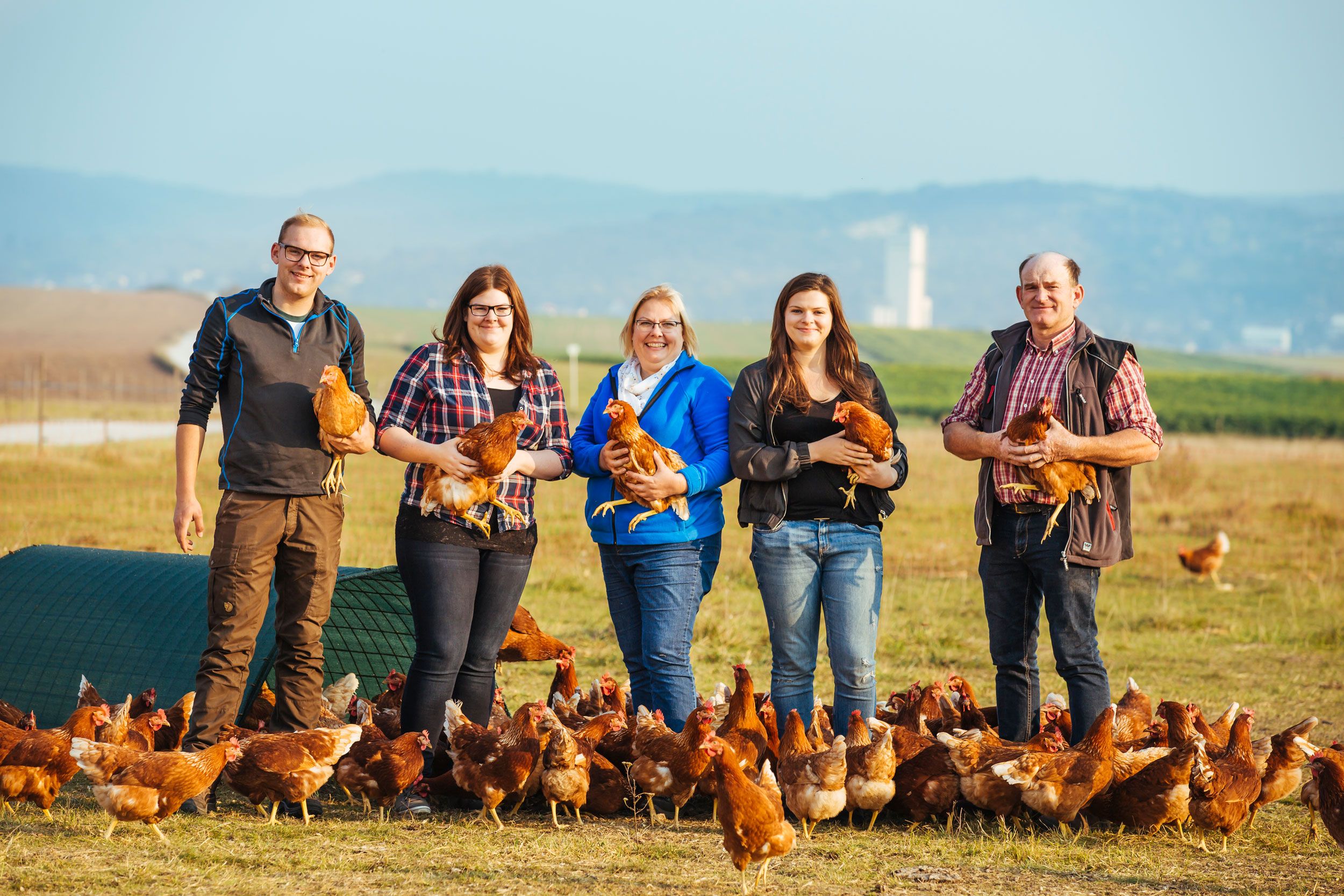 A family stands in a field with chickens and holds some in their arms.