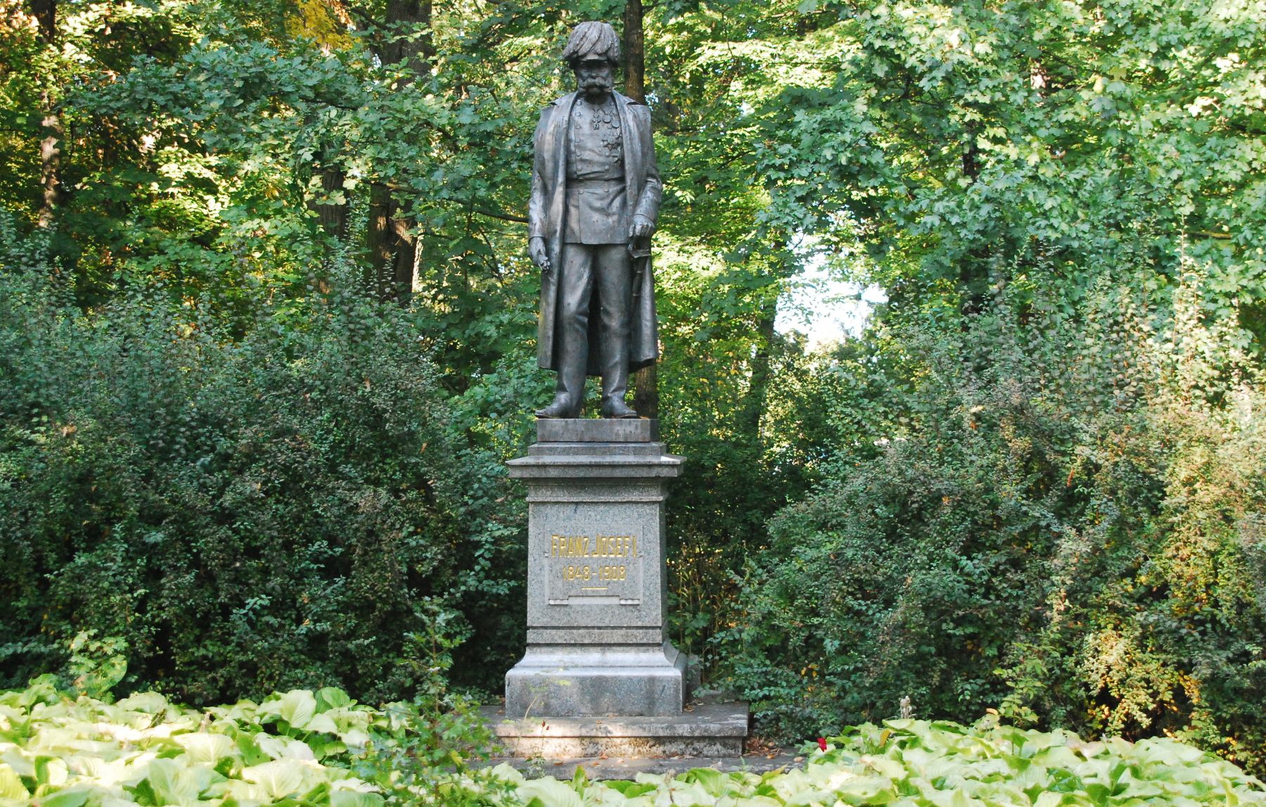 Statue of Emperor Franz Joseph I in a park, surrounded by trees and shrubs.