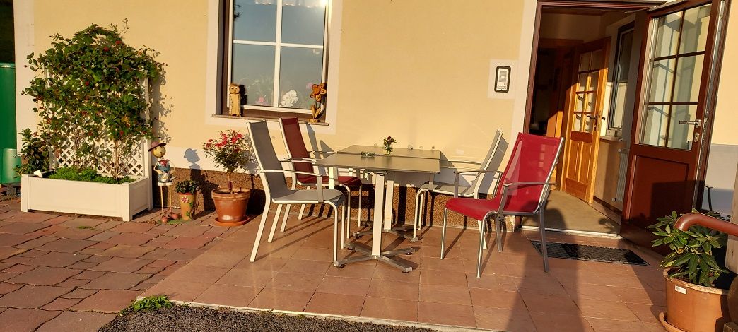 Terrace with table and chairs in front of a building, surrounded by plants.