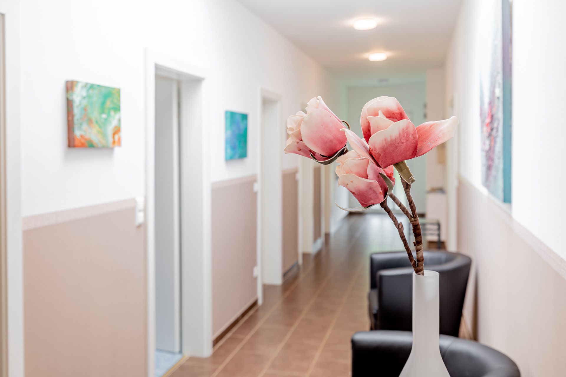 A bright hallway with white walls, decorated with works of art and a vase of pink flowers in the foreground.