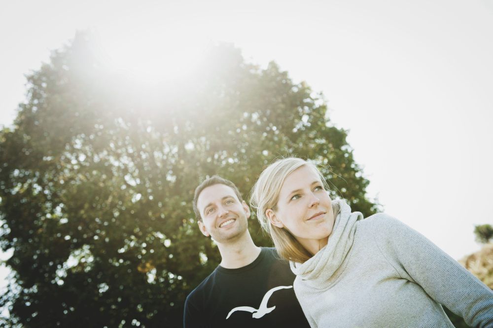 A man and a woman are standing outside in front of a tree, the sun shining through the leaves.