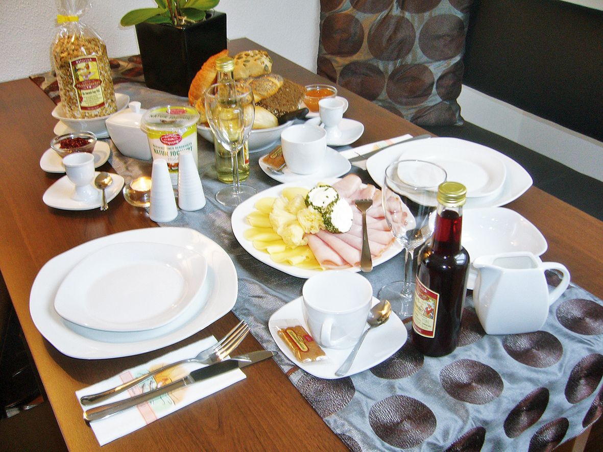 Breakfast table with crockery, cold cuts, bread, muesli and drinks.