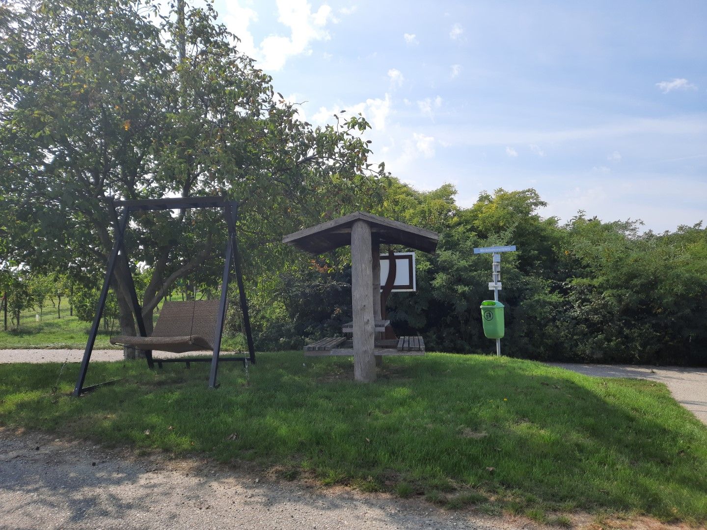 A rest area with a swing and a covered seating area next to a tree and a garbage can on a meadow.