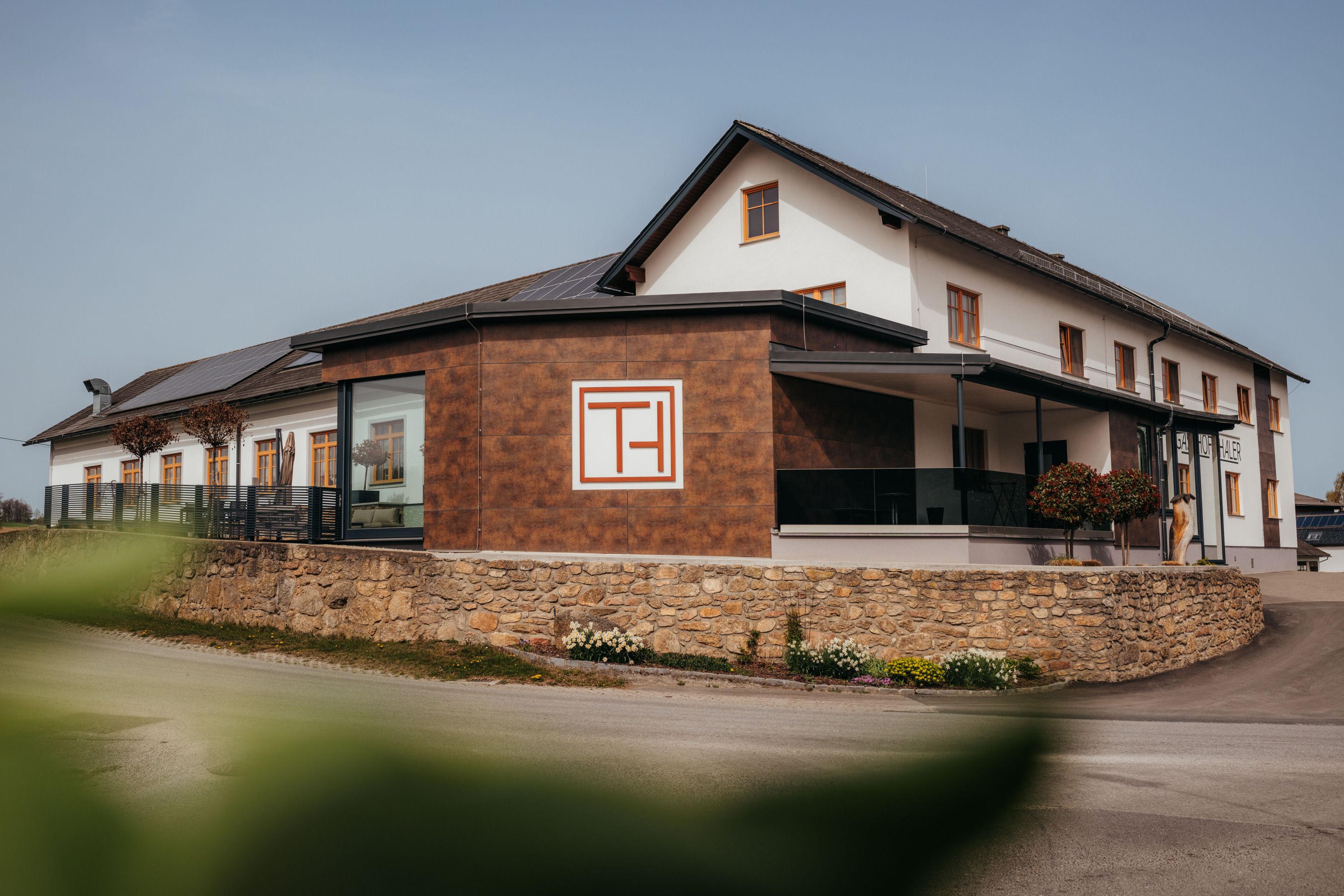 A modern building with the inscription 'Gasthof Thaler', surrounded by a stone wall and plants.