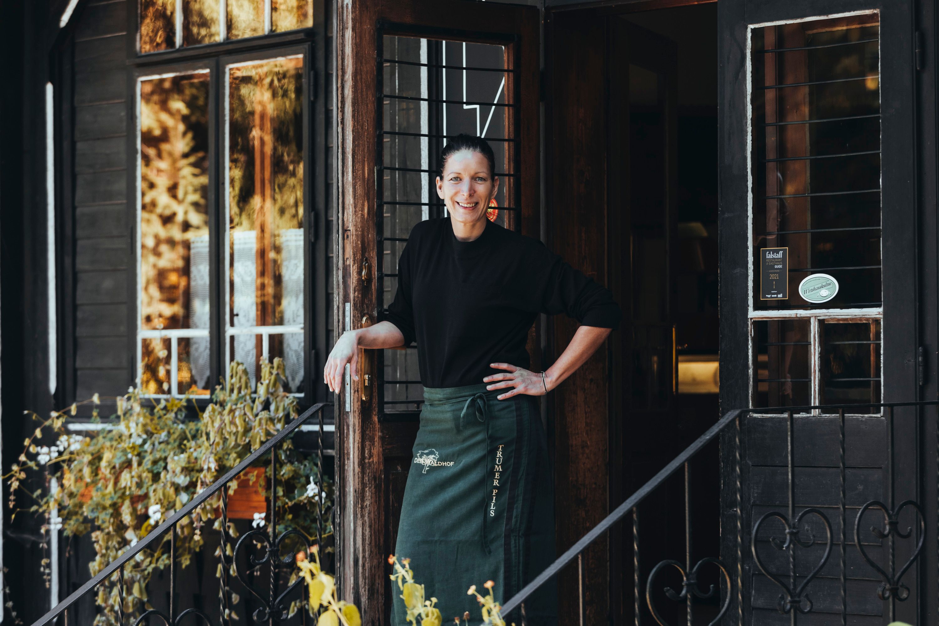A woman stands smiling in the doorway of a rustic building with plants in the foreground.