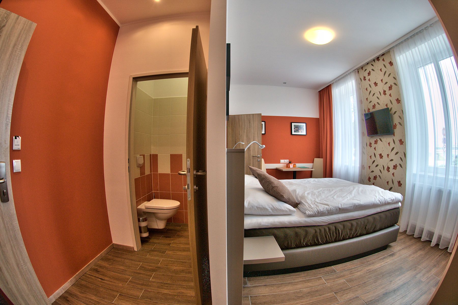 A modern guest room with double bed, desk and TV. Next to it a small bathroom with toilet.