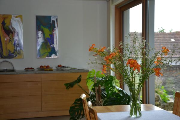 A breakfast room with a table on which there is a bouquet of flowers. In the background are works of art on the wall and a sideboard with fruit.