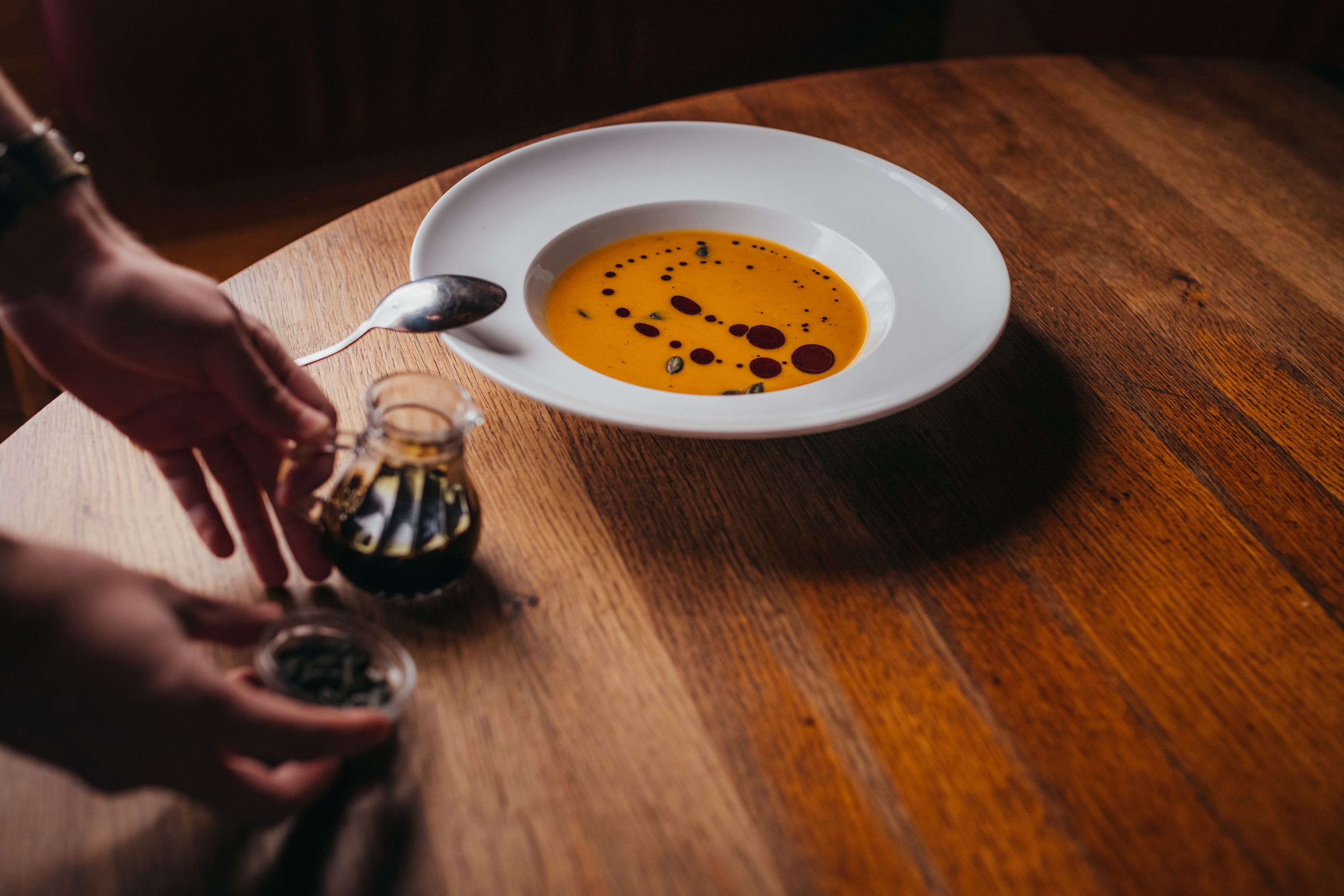 A plate of pumpkin soup with oil on a wooden table, hands with spices next to it.