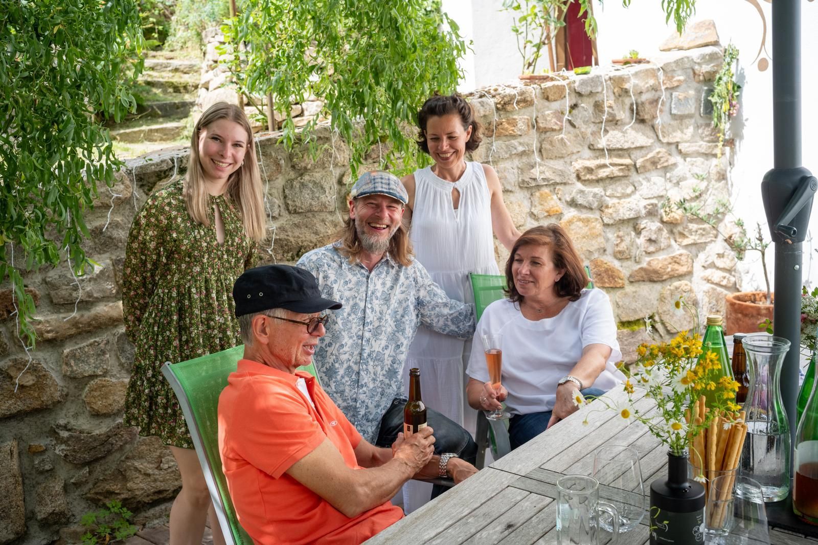 Group of five people sitting and standing at an outdoor table, surrounded by plants and a stone wall.