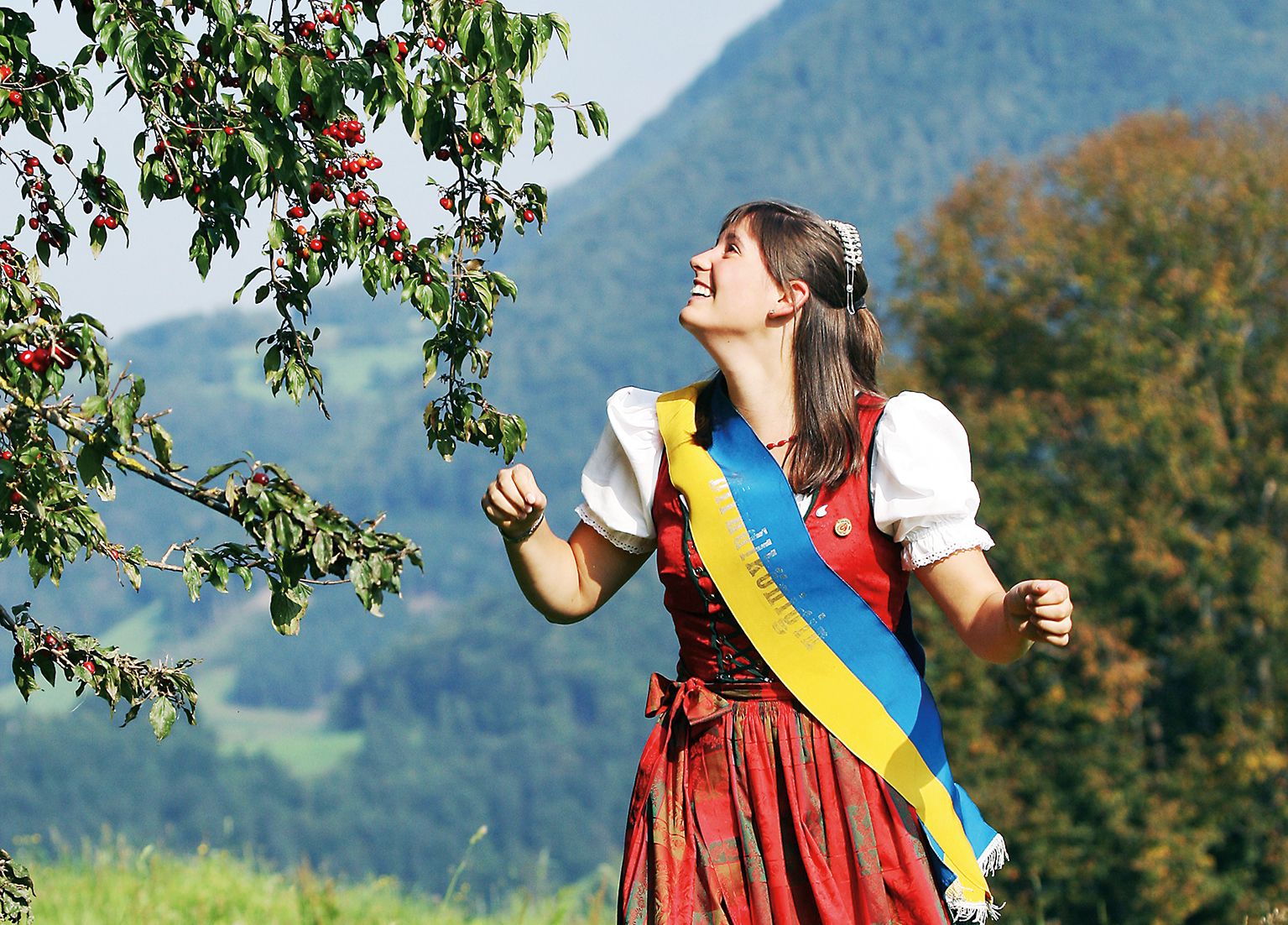 Woman in traditional costume stands under a tree with red fruits in a rural landscape.