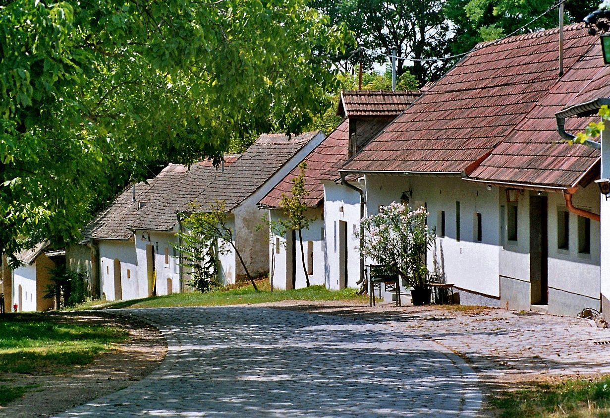 A cobbled street leads past a row of traditional white houses with red roofs, surrounded by green trees.