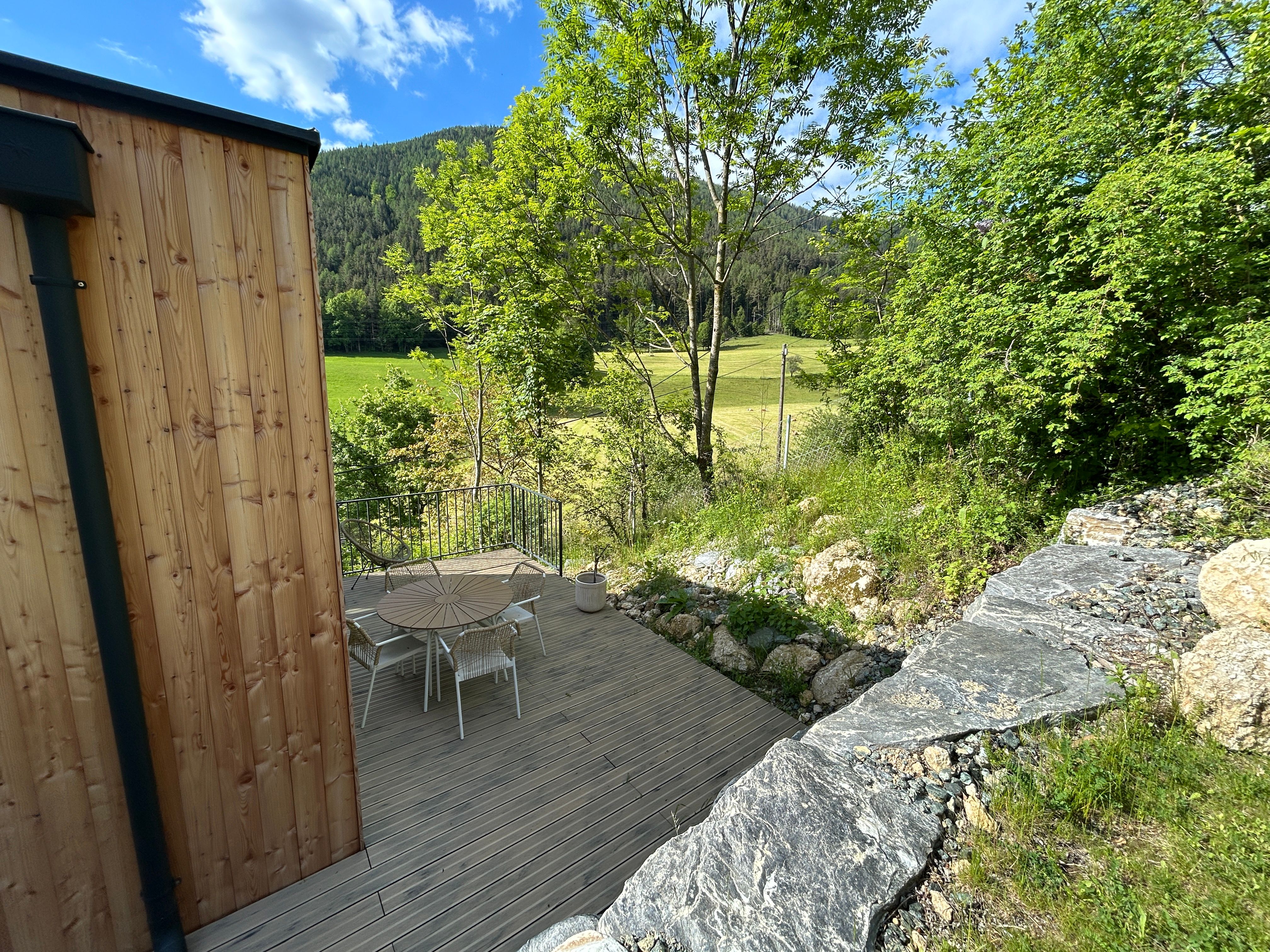 Terrace with wooden table and chairs, surrounded by trees and meadow, with a view of wooded hills.
