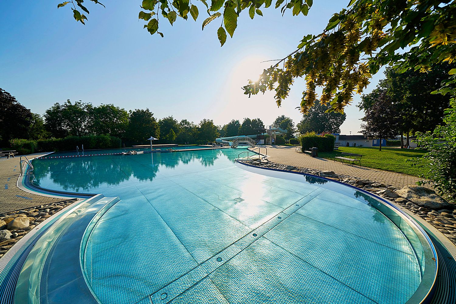 An outdoor pool with clear water, surrounded by trees and a sunny sky.